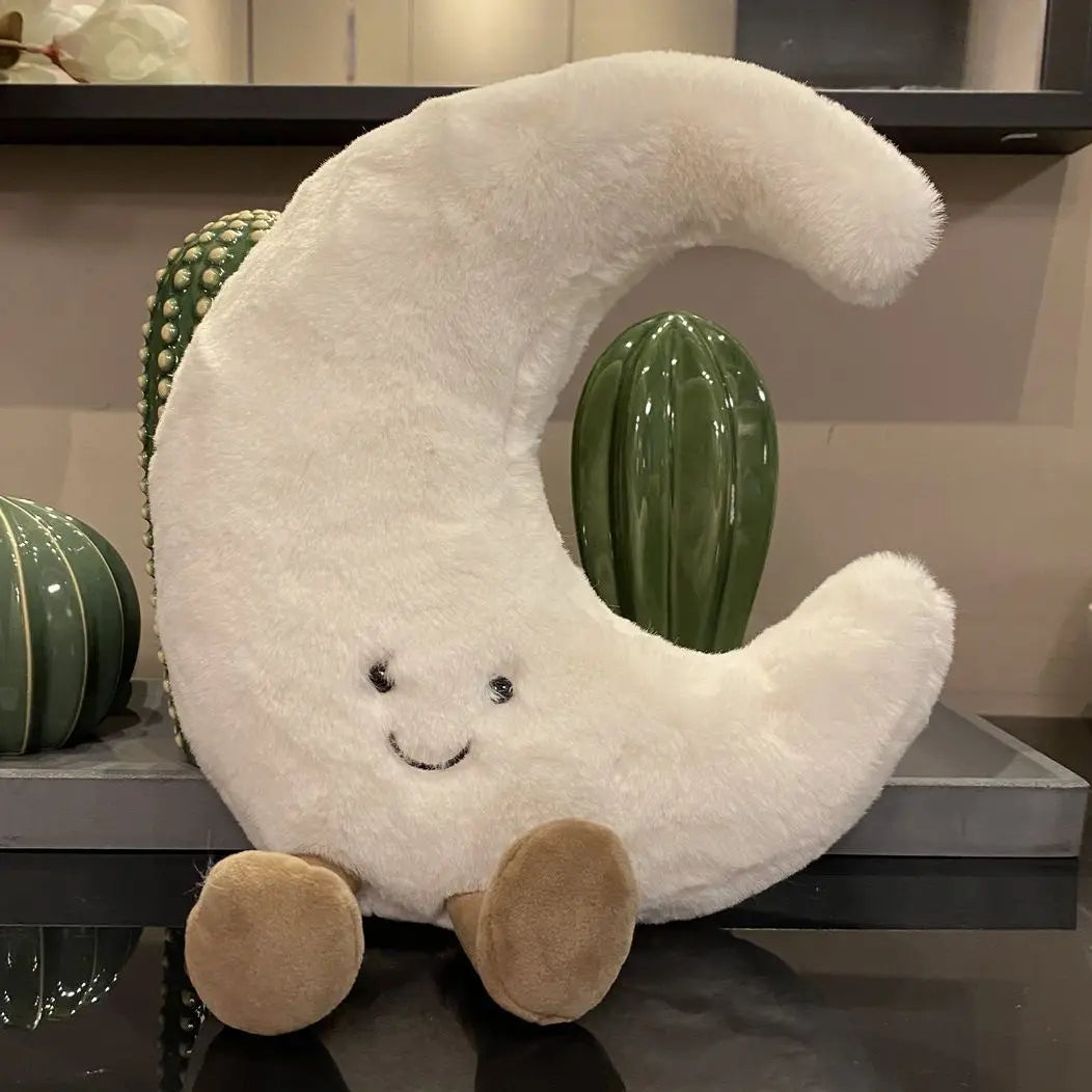 A white crescent moon plushie with a smiling face and tan feet is displayed next to a green cactus.