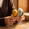 A pair of handmade crochet dinosaur keychains, one yellow and one green, are held by a person on a wooden table.