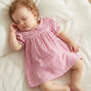 A baby girl wearing a pink and white gingham A-line dress with a Peter Pan collar lying down on a white bed.
