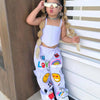 A young girl wearing a white suspender top and long purple pants with colorful graphics and sunglasses.