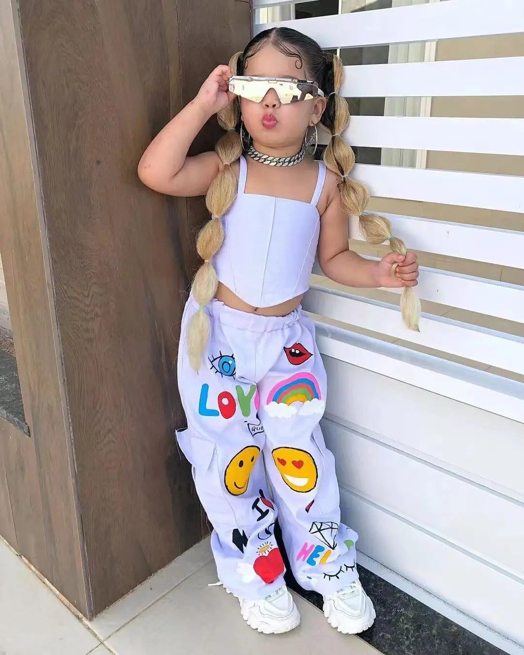 A young girl wearing a white suspender top and long purple pants with colorful graphics and sunglasses.
