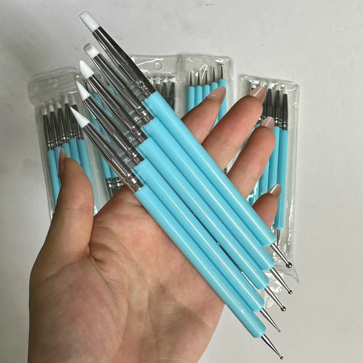 A hand holds a clear plastic container of a 5-piece double-ended silicone dotting pen set with light blue handles and silver 