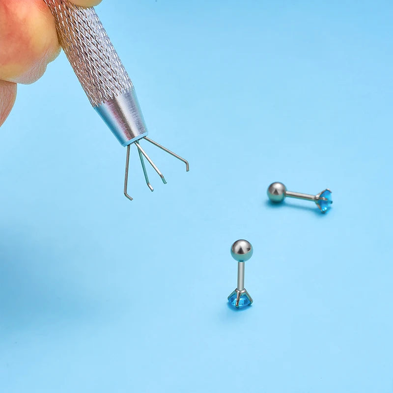 A silver precision tool with multiple prongs is shown picking up a small blue bead or jewelry piece.