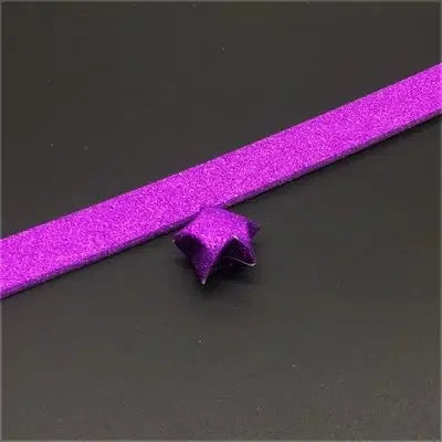 A purple glitter origami star paper strip is shown lying on a dark surface.