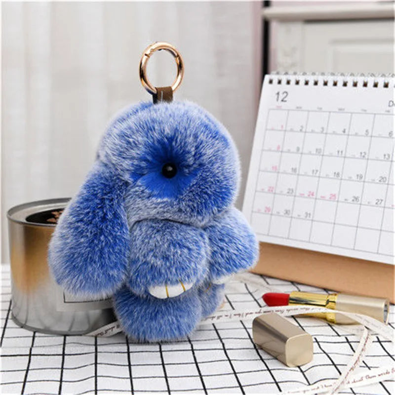 A fluffy blue plush bunny keychain is displayed on a white checkered surface next to a metal tin.