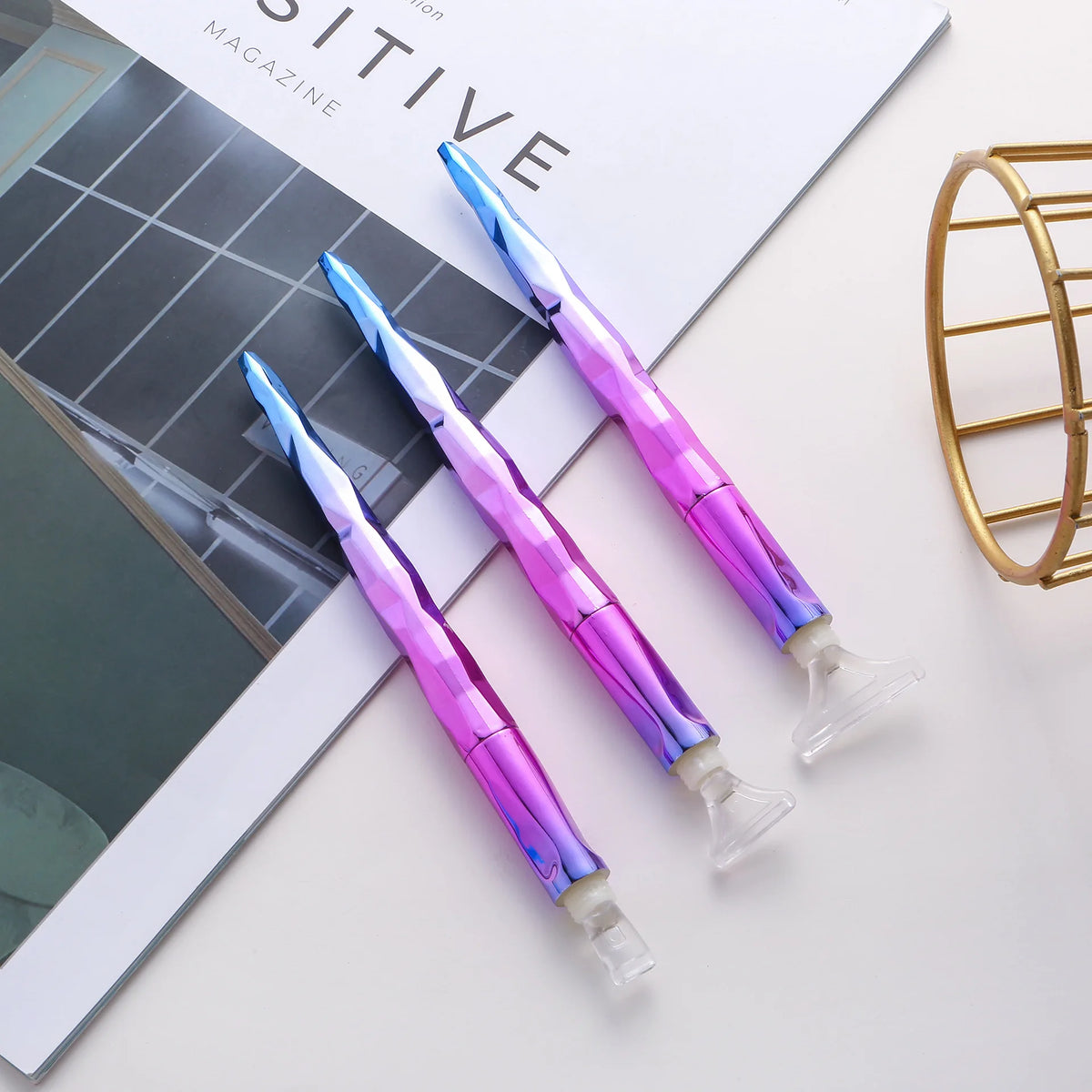 Three iridescent purple diamond painting pens with roller tools displayed on a white surface next to a magazine.