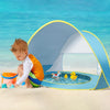 Kleinkind sitzt in einem blau-gelben Sofortaufstell-Strandzelt an einem Sandstrand mit klarem blauem Wasser im Hintergrund.