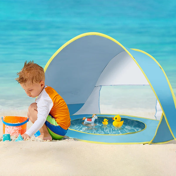 A toddler sits inside a blue and yellow instant pop-up beach tent on a sandy beach with clear blue water in the background.