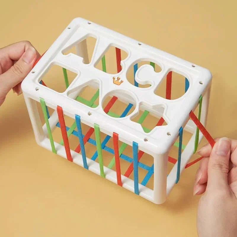 A white Montessori shape sorting cube with letter cutouts and elastic bands is being held by a hand.