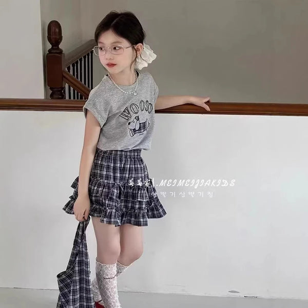 A young girl wearing a plaid skirt and gray shirt with a cartoon print, standing in a room.