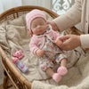 A newborn baby doll with pink knitted clothing is cradled in a wicker basket on a beige blanket.