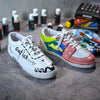A white sneaker with black lettering and dripping paint is displayed on a gray fabric surface.
