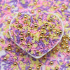 A heart-shaped dish filled with colorful polymer clay slices in pink, purple, and yellow mixed with gold glitter particles.