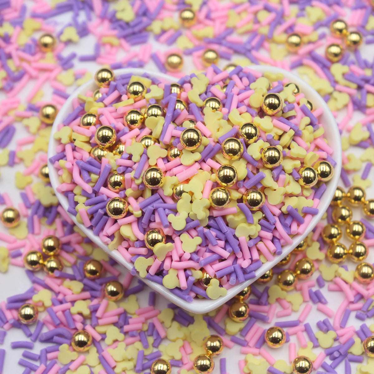 A heart-shaped dish filled with colorful polymer clay slices in pink, purple, and yellow mixed with gold glitter particles.