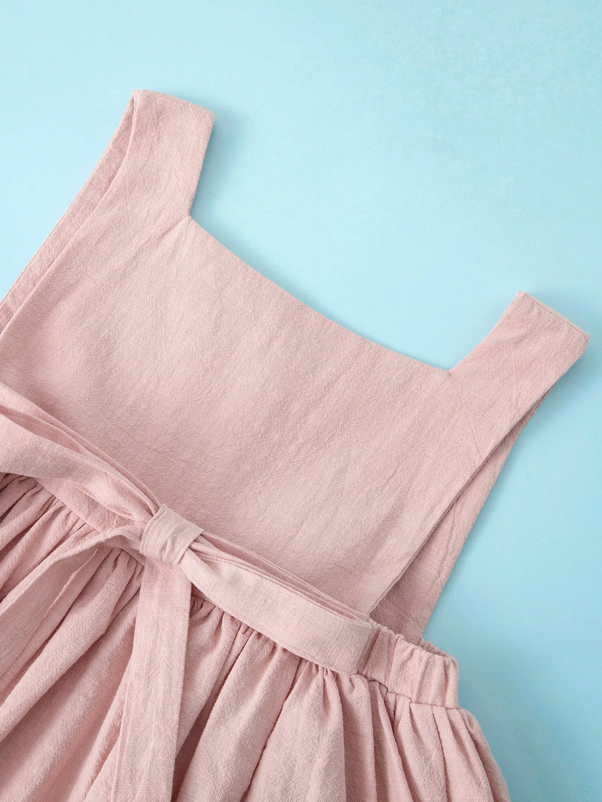 A light pink summer cotton linen jumper dress for a toddler girl, shown flat lay against a light blue background.