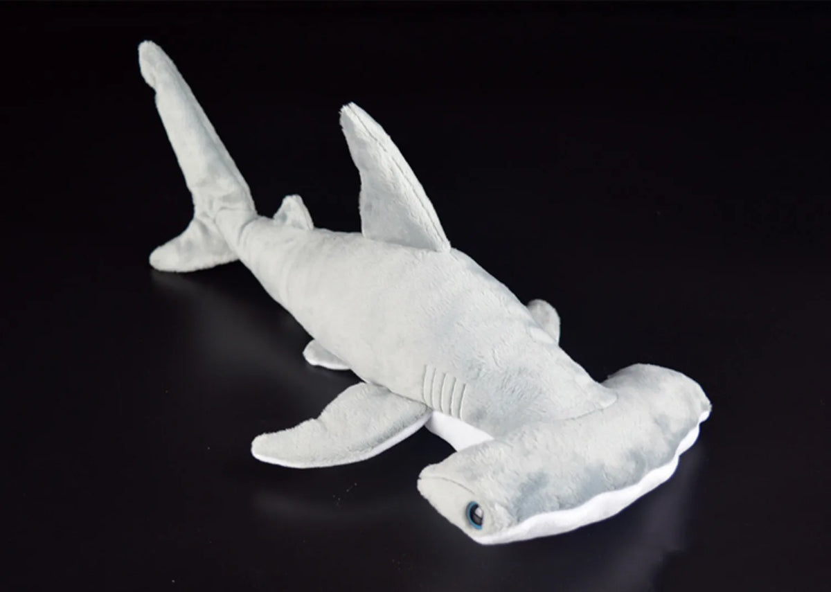 A white super soft hammerhead shark plush toy viewed from the side against a dark background.
