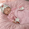 A newborn baby doll wearing a floral cap and pink outfit is lying on a fluffy pink surface.