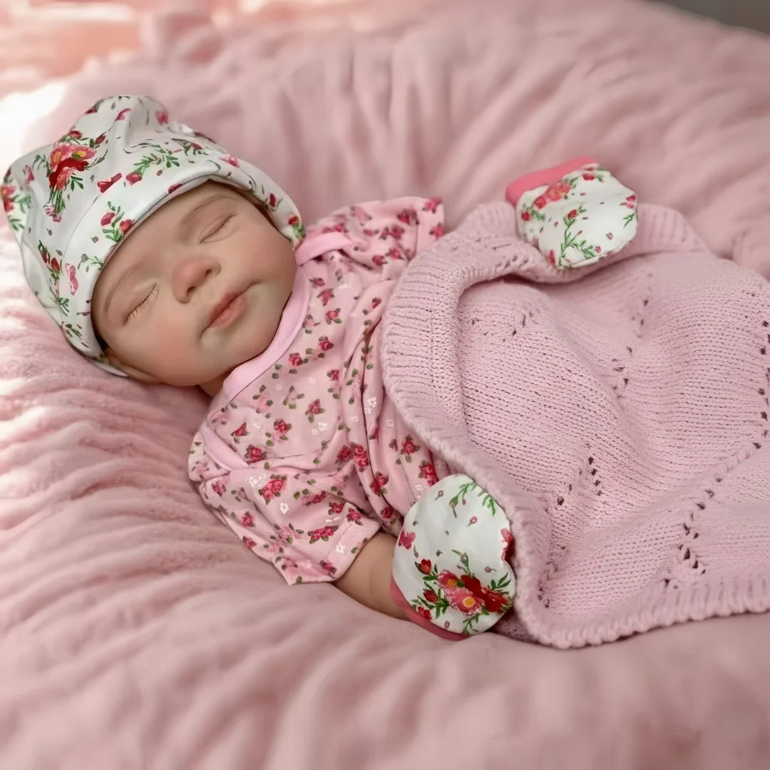 A newborn baby doll wearing a floral cap and pink outfit is lying on a fluffy pink surface.