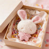 A white plush bunny keychain with pink embroidered ears is displayed in a light-colored box lined with shredded paper.