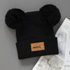 Black knit baby beanie hat with large black pom-poms and a small brown mini tag