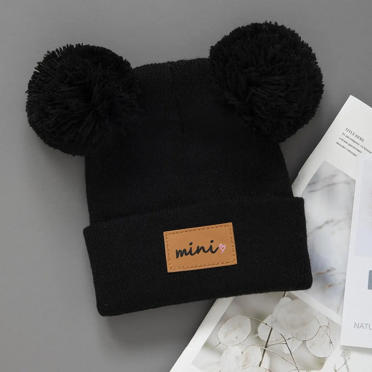 Black knit baby beanie hat with large black pom-poms and a small brown mini tag