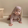 A baby wearing a light pink rabbit romper set with a matching hat and socks, sitting on a light surface.