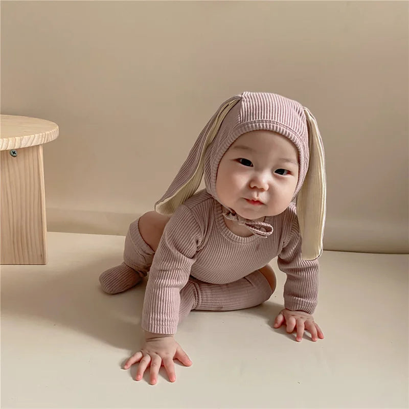 A baby wearing a light pink rabbit romper set with a matching hat and socks, sitting on a light surface.