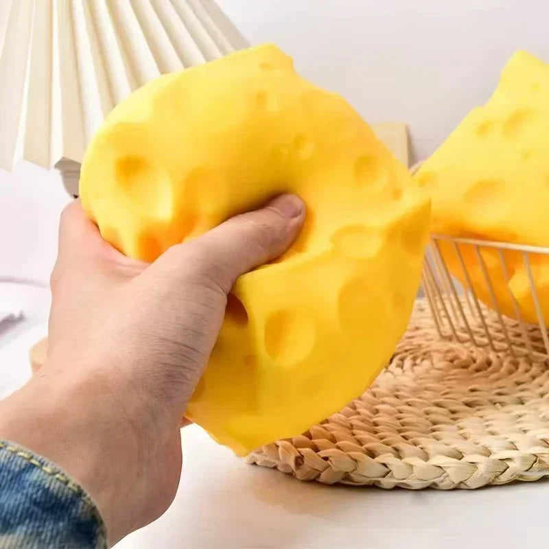 A hand is squeezing a bright yellow cheese wedge squishy stress relief toy on a woven mat.