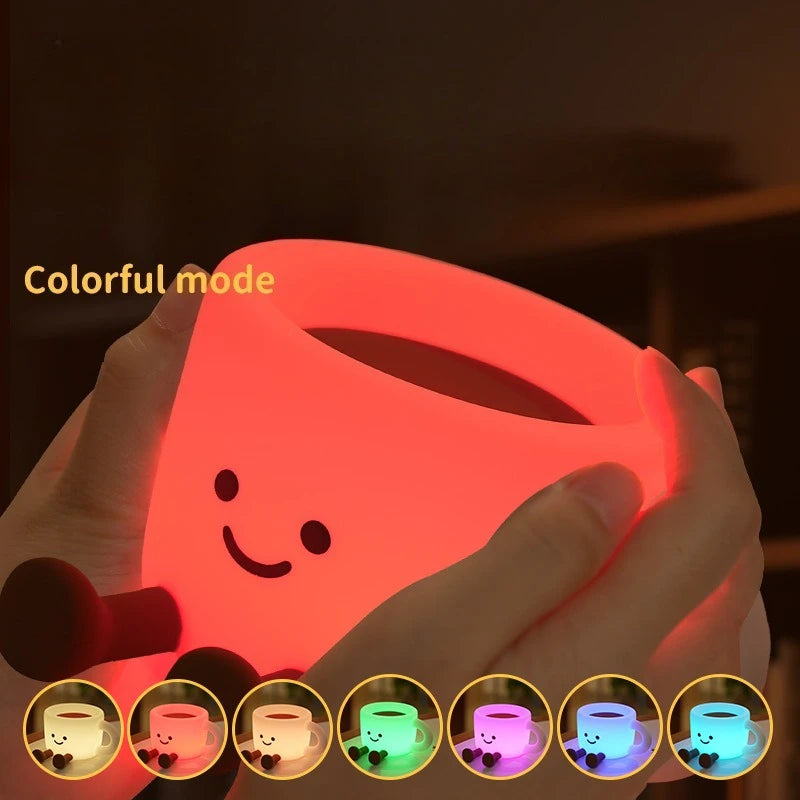 Hands hold a colorful coffee cup shaped night light illuminating a smiling face and various colors.