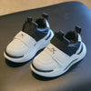 A pair of white and black kids' everyday sneakers with easy velcro straps resting on a dark surface.
