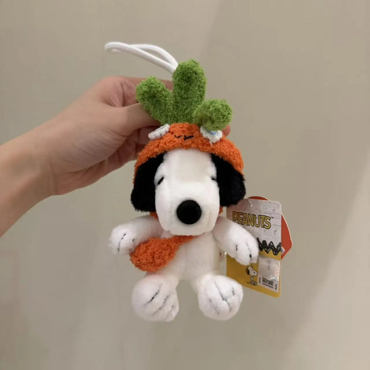 A white Snoopy plush charm with green leaves and an orange hat is being held against a light background.