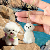 A clear, dog-shaped pet hair keepsake keychain is held by a hand against a backdrop of rocks and blue water.