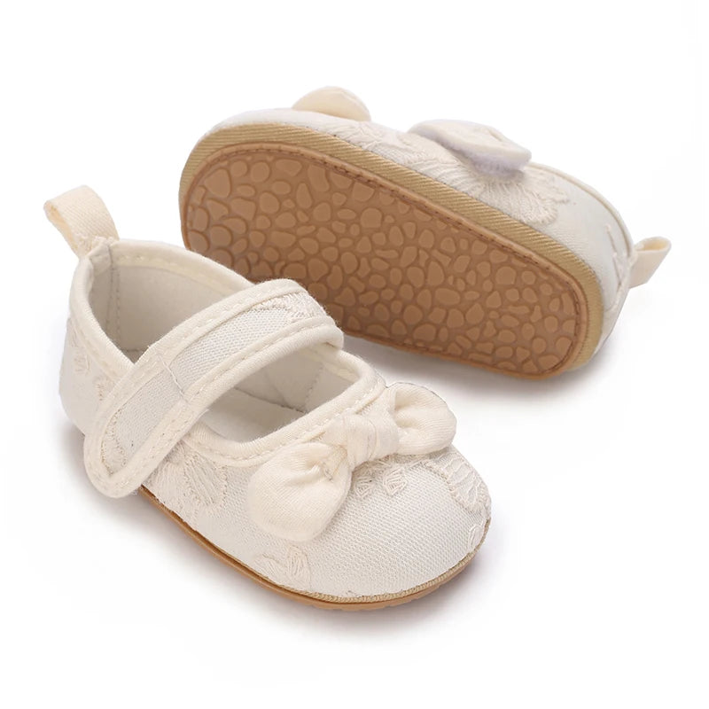 A pair of light beige lace-trimmed baby girl moccasin shoes with a bow, shown from the top and side.