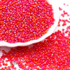 A close-up view of bright red glossy glass seed beads scattered on a white dish and spilled onto a surface.