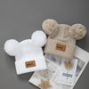 A beige knit baby beanie hat with fluffy pom-poms and a small brown tag is shown on a light gray background.