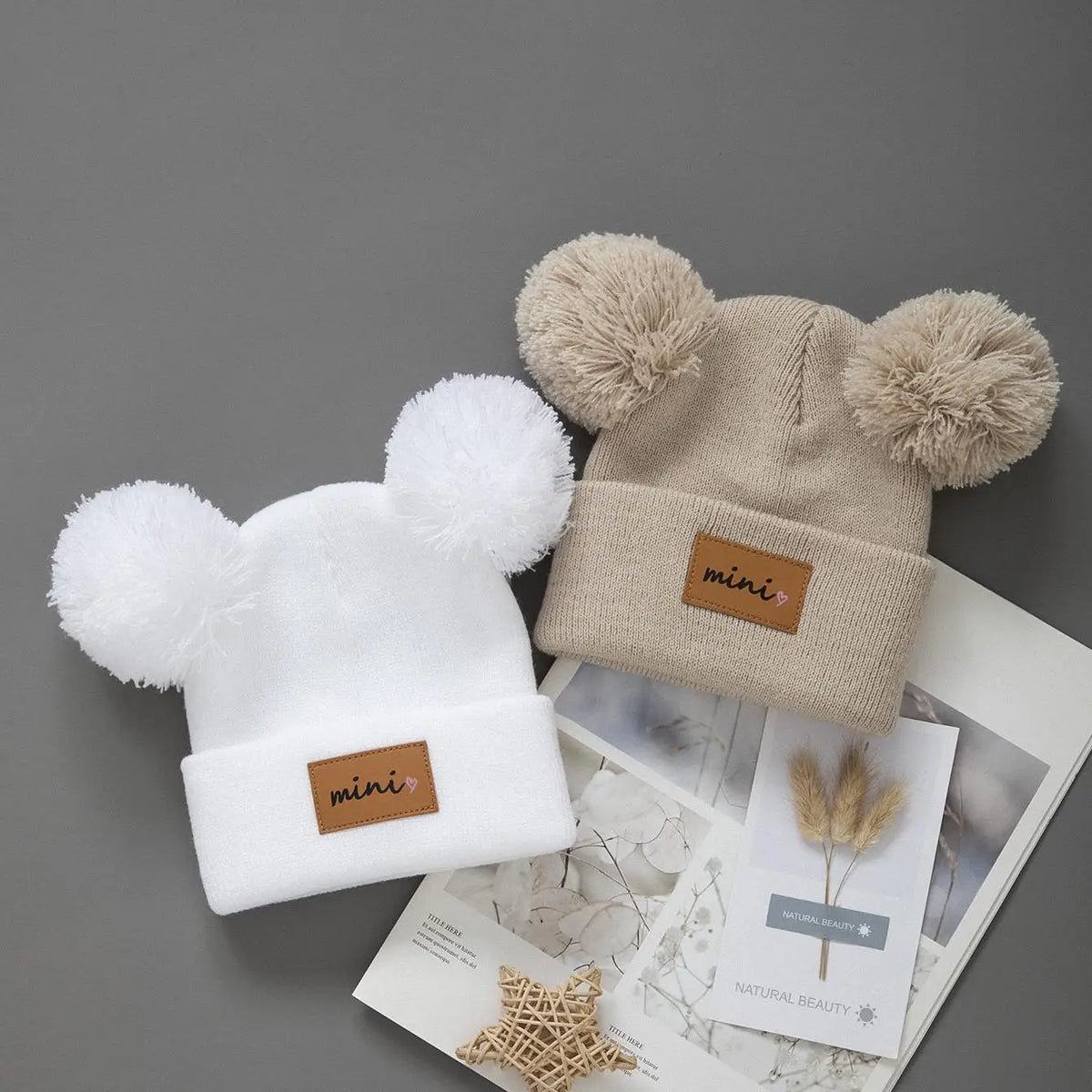 A beige knit baby beanie hat with fluffy pom-poms and a small brown tag is shown on a light gray background.