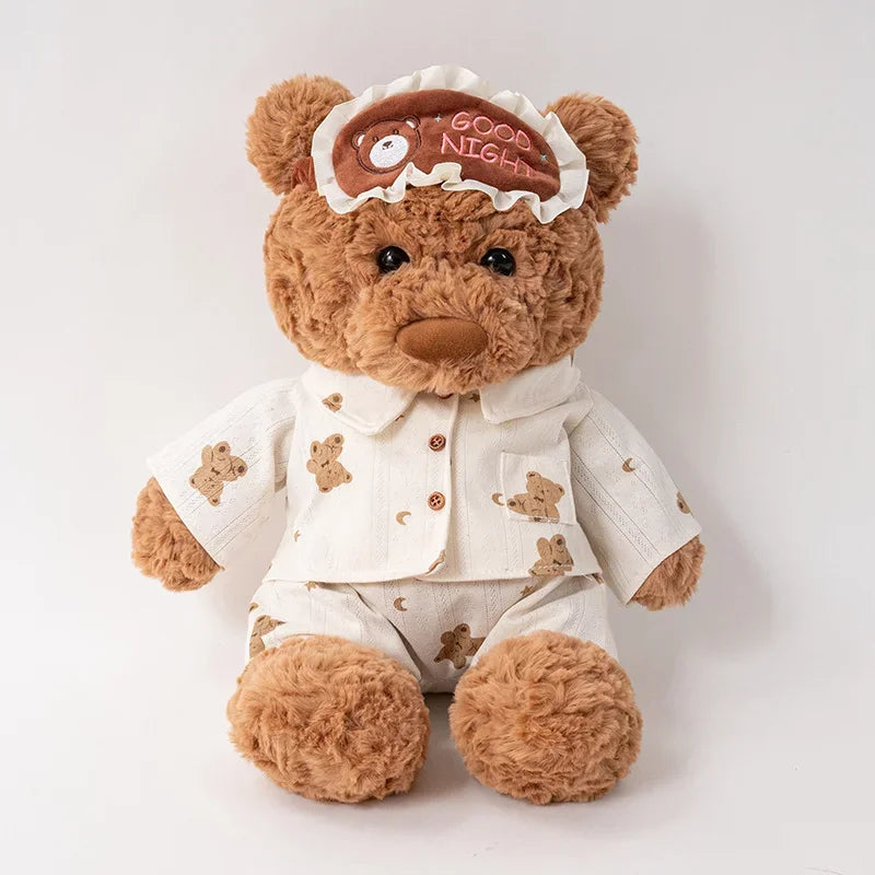 A fluffy brown teddy bear plush toy wearing a white collared shirt and pants with an eye mask and birthday cake decoration.