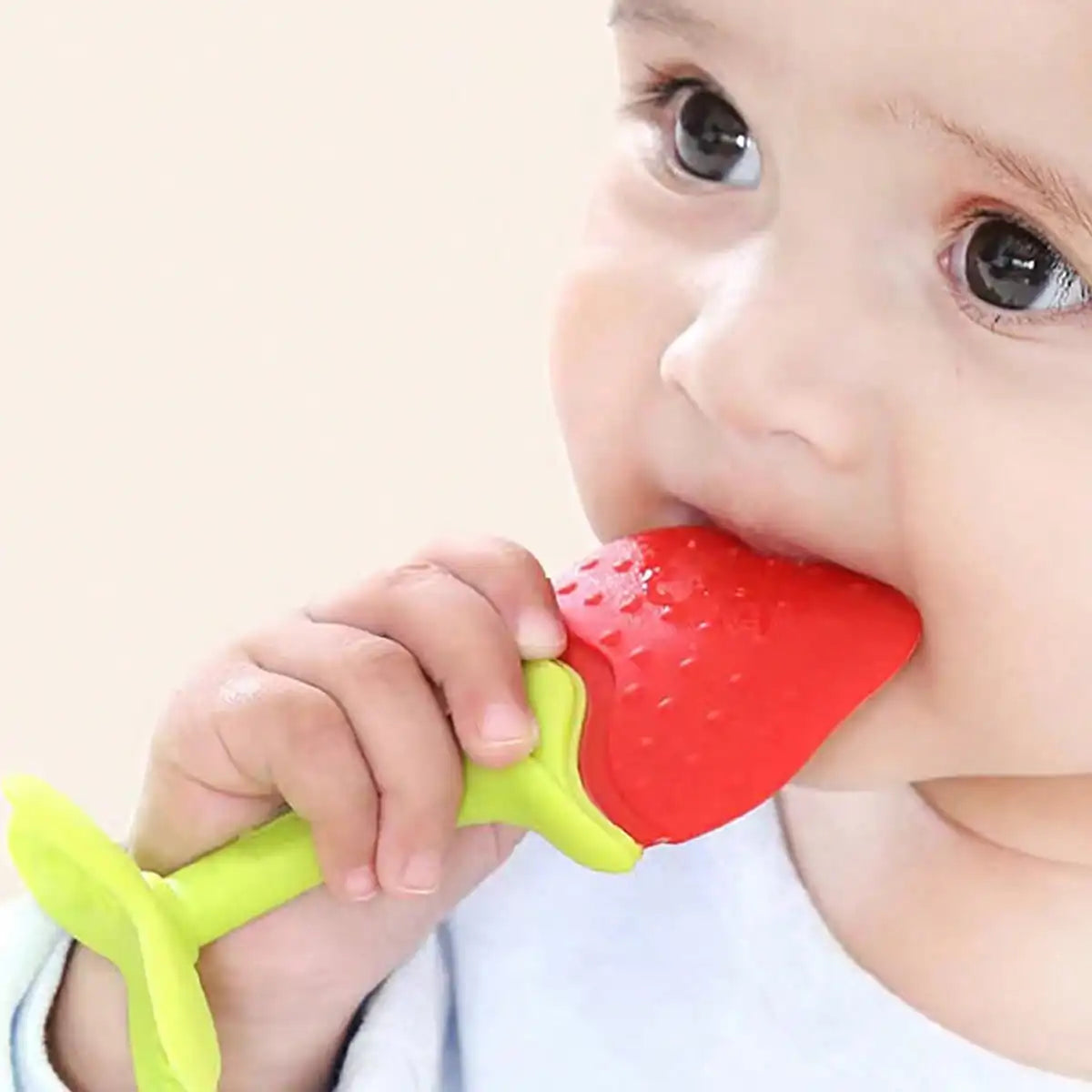 A baby is holding a bright red silicone fruit-shaped teether with a yellow handle.