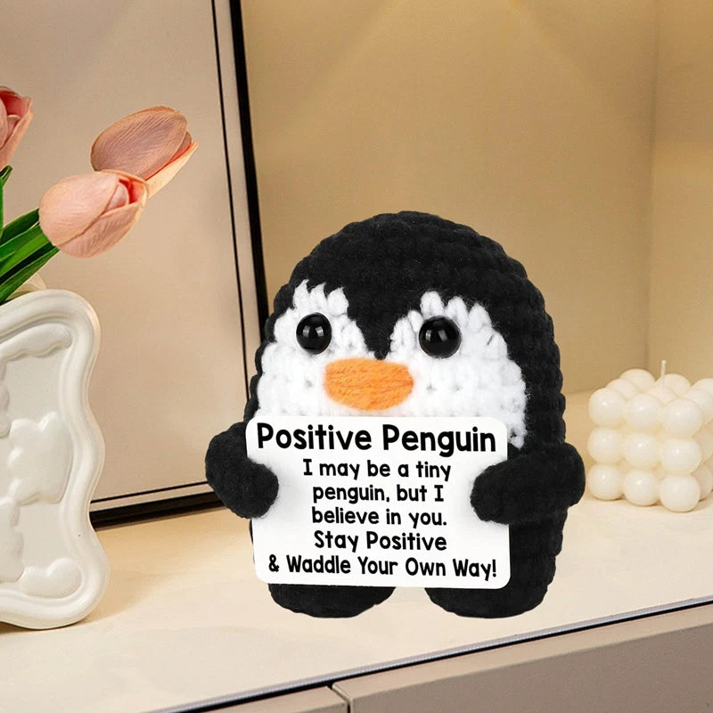 A handmade crochet penguin doll with a black and white pattern and a sign that reads I may be a tiny penguin but I believe in