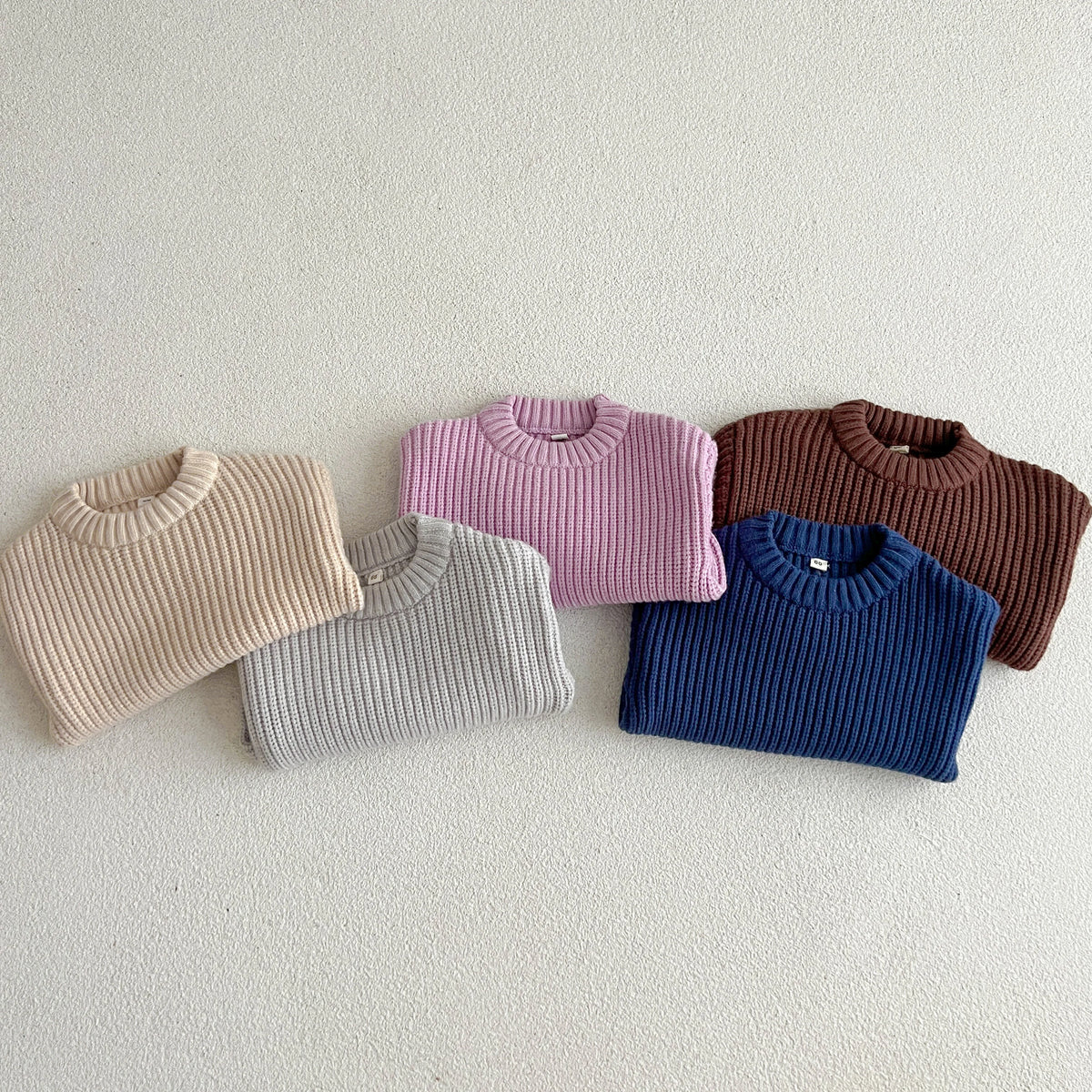 A row of five knitted baby pullovers in pastel and earthy tones laid flat on a light background.