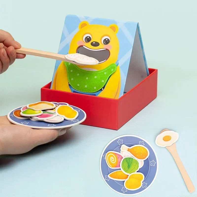 A yellow bear toy in a blue box is being fed with a wooden spoon, accompanied by a plate of colorful food items.