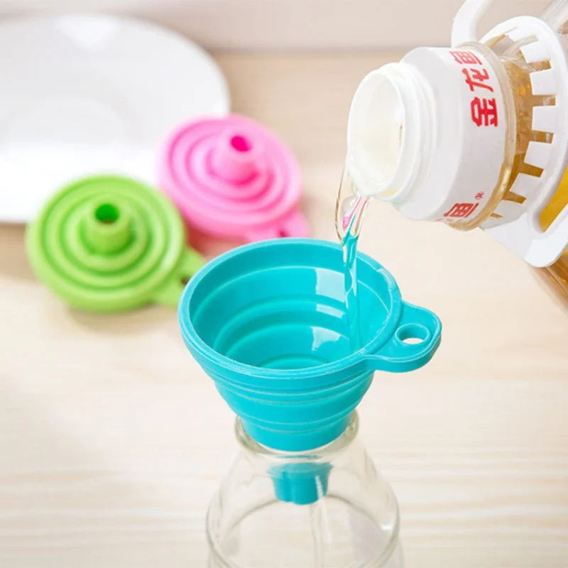 A clear glass bottle is being filled with oil using a white funnel, next to colorful silicone funnels.