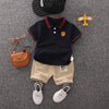 A navy blue short sleeve t-shirt and beige shorts set for a boy, with a small bear charm and a yellow airplane decoration.
