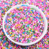A white bowl filled with colorful polymer clay slice sprinkles for slime and DIY crafts scattered on a pink surface.
