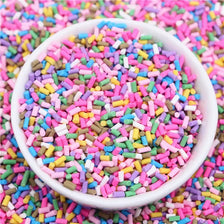 A white bowl filled with colorful polymer clay slice sprinkles for slime and DIY crafts scattered on a pink surface.