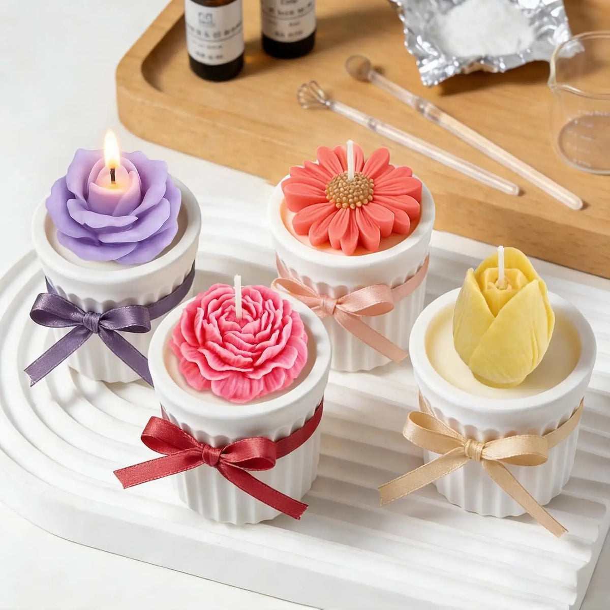 A set of four silicone flower molds in various shades of pink, purple, and yellow with lit candles displayed on a white tray.