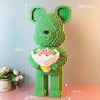 A green bear figure built from micro bricks, holding a pink flower bouquet, standing on a pink surface.