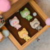 A wooden box contains several small, cute cat-shaped figurines in various pastel colors like green, pink, and yellow.