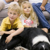 Two children are sitting on a couch with a black and white dog lying in front of them.