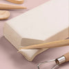 A block of white air dry modeling clay is shown with wooden tools and shaping tools on a pink surface.
